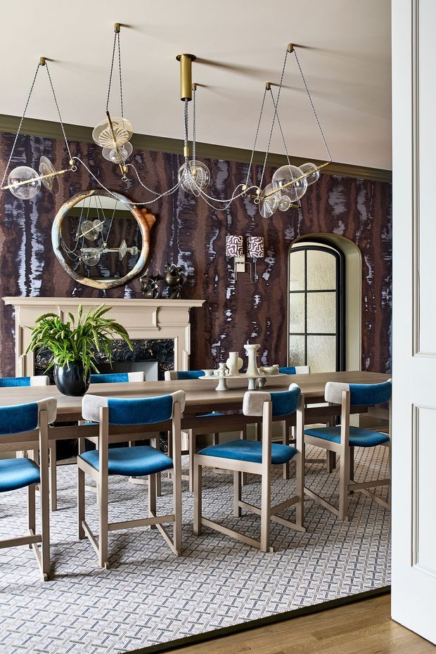 A richly styled dining room viewed through an open white paneled doorway. A long dark timber dining table is surrounded by light timber-framed chairs upholstered in vivid blue velvet. The room features a pale traditional fireplace mantel, ornate round mirror, dark patterned feature wallpaper, a tall arched black-framed glazed door or window, decorative candlesticks, greenery, and sculptural brass pendant lighting suspended from the ceiling. A geometric patterned carpet covers most of the floor, with timber flooring visible at the threshold. The scheme is dramatic, refined, and eclectic, combining traditional architectural detailing with contemporary furnishings and strong blue and dark brown tones.