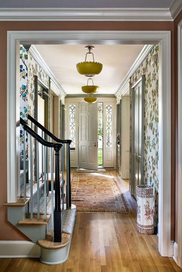 A bright, richly detailed residential entry hall viewed from an adjoining room. The space features a timber staircase with dark turned handrail and pale balusters, warm timber flooring, patterned runner rugs, floral botanical wallpaper, substantial white decorative mouldings and cornices, grey-green doors and trim, and a white front door with narrow decorative glass sidelights. Three distinctive mustard pendant lights run along the hallway, while a patterned cylindrical umbrella stand adds a decorative accent.