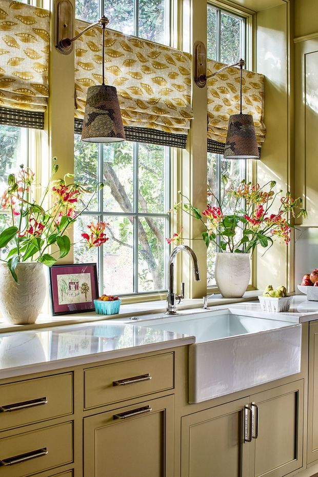 Bright, richly detailed kitchen vignette centred on a large farmhouse-style apron-front sink beneath tall multi-pane windows. Pale olive-green painted cabinetry with framed doors and drawers, pale stone countertops, polished chrome tapware, patterned botanical Roman blinds, pendant lights, and abundant floral arrangements create a warm traditional character.