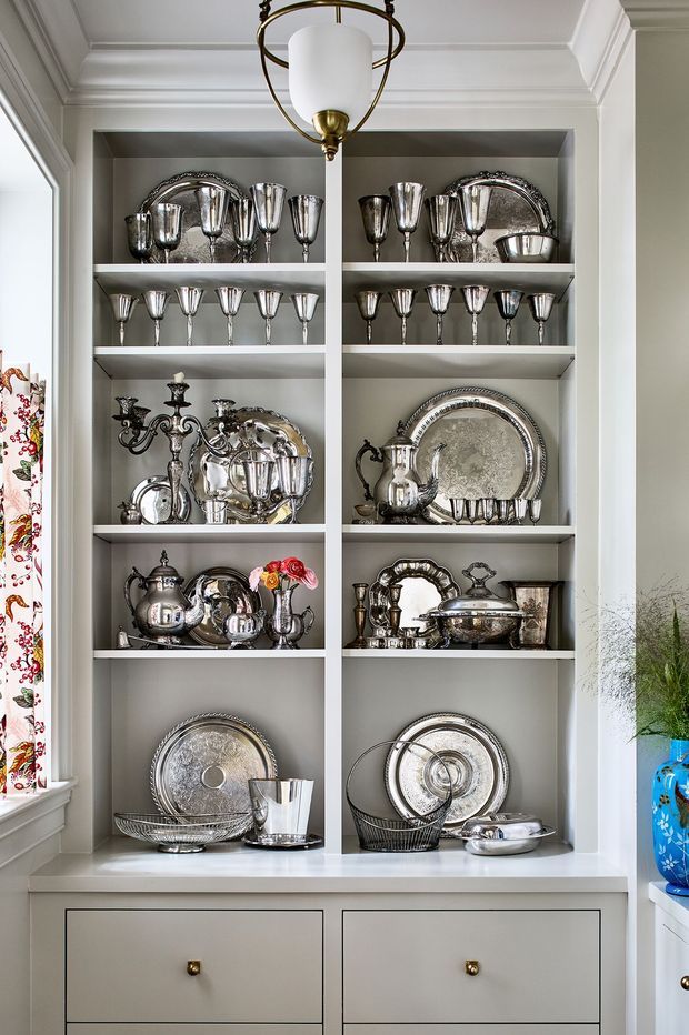 A built-in white display and storage cabinet with open shelving, drawers, and ornate silver servingware, trays, goblets, and decorative pieces arranged throughout. The cabinet is set within a bright traditional interior, with detailed crown moulding, a brass-and-white pendant light, and glimpses of floral fabric and a blue decorative vessel at the sides.