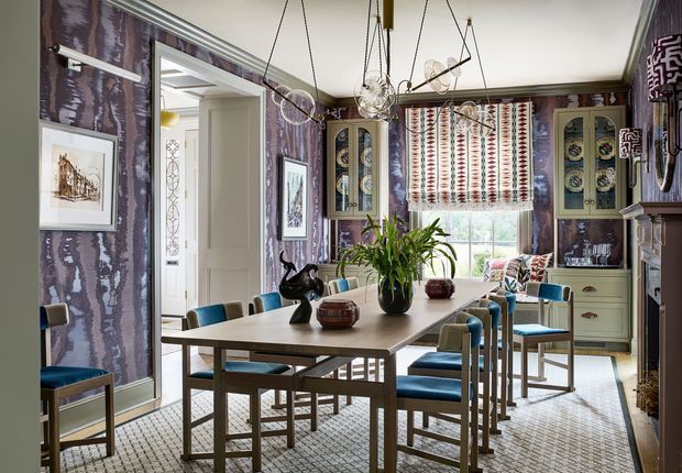 Eclectic dining room with a long rectangular timber dining table surrounded by blue upholstered chairs, patterned wallpaper, built-in display cabinetry, a large window with patterned Roman blind, and multiple decorative pendant lights. The room has a richly layered, colourful and highly detailed interior with a classic architectural shell and contemporary furnishings.