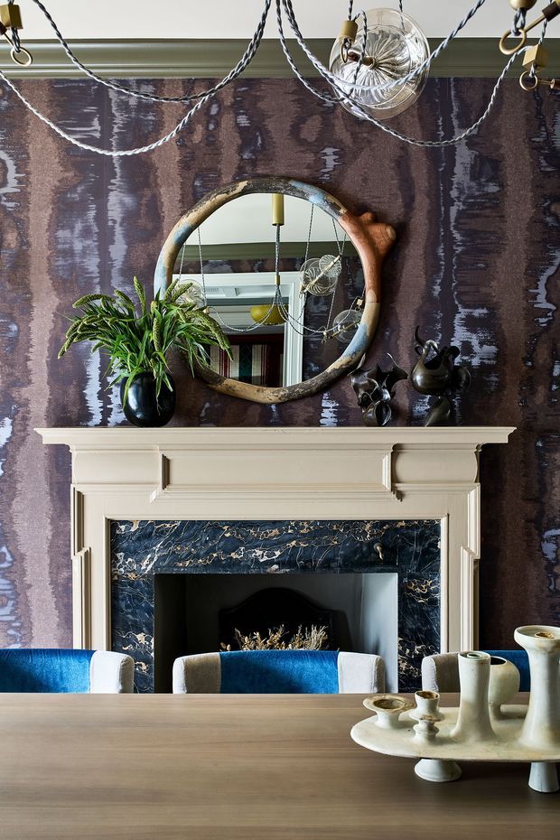 A richly layered interior vignette centred on a classical mantelpiece with cream painted timber detailing and a dark blue-black marble fireplace surround with prominent veining. The wall is covered in a dramatic dark mauve and grey patterned finish. An organic sculptural round mirror, hanging decorative lights, leafy plant, dark ceramic objects, and vivid blue upholstered dining chairs surround a large timber dining table in the foreground. The overall composition is eclectic, refined, and highly decorative.