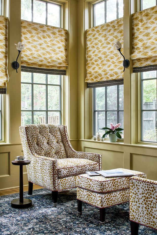 A warmly styled sitting area in a bay of tall, multi-pane windows. Olive-beige painted walls and window trim frame patterned Roman blinds, while two upholstered armchairs and a matching ottoman feature a bold cream-and-mustard spotted fabric. A dark patterned area rug, small round side table, and potted orchid complete the refined, eclectic interior.