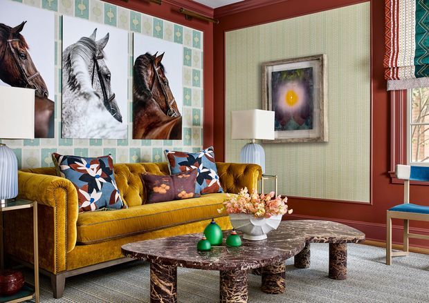Eclectic, richly layered living room featuring a mustard tufted sofa, patterned botanical cushions, horse artwork, a dark marble coffee table, striped carpet, red-painted trim, and green patterned wallcovering. Decorative Roman blinds and a blue accent chair add further colour and visual interest.