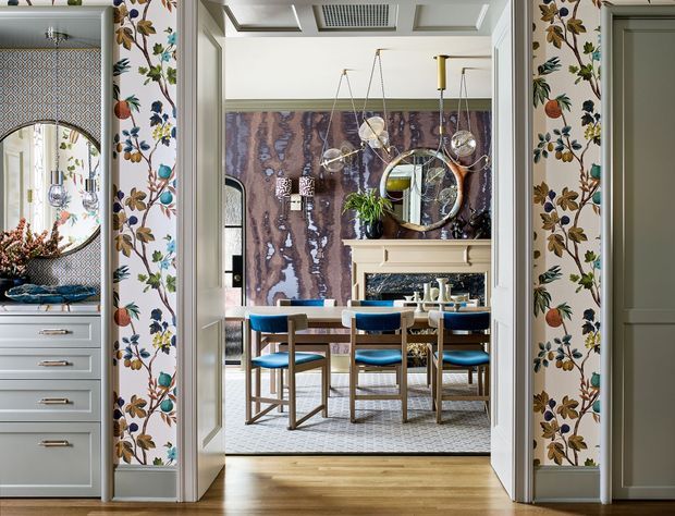 A richly decorated transitional dining room viewed through an open doorway, featuring a rectangular dining table with blue upholstered chairs, a sculptural multi-light pendant, ornate floral wallpaper on the surrounding walls, a dramatic marbled feature wall, a fireplace or mantel with mirror above, patterned carpet, and light timber flooring in the foreground. Built-in cabinetry and a decorative oval mirror are visible in the adjacent space.