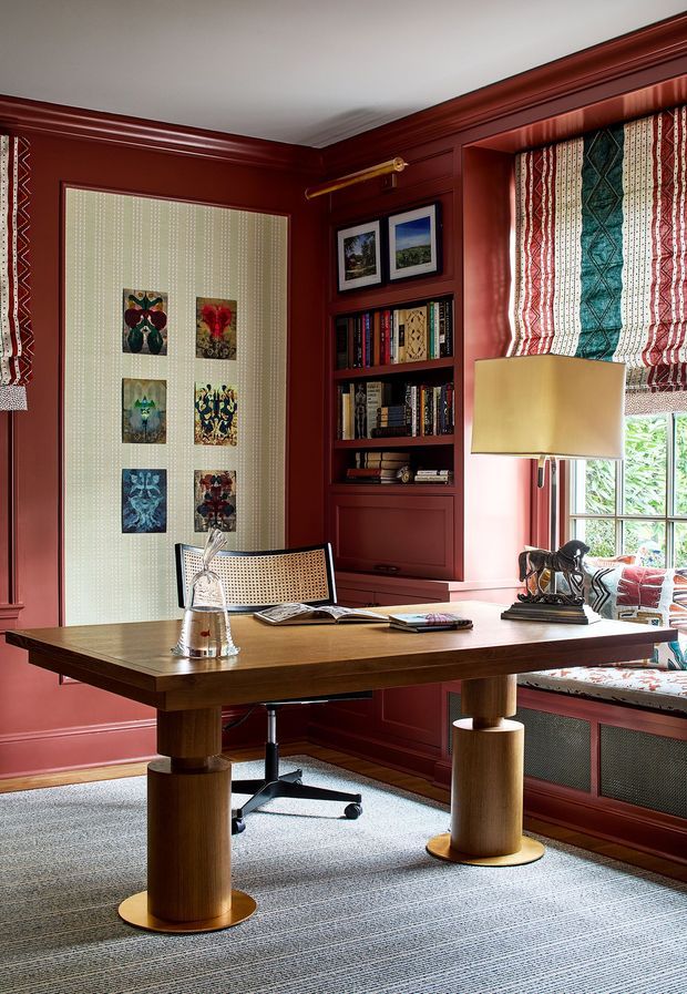 A richly detailed home office with deep red painted walls, extensive matching timber trim, and a built-in bookcase. A large wood desk with cylindrical pedestal legs occupies the foreground, paired with a cane-back office chair. Patterned fabric blinds frame the window and coordinate with a cushioned window seat. The room includes framed artwork, books, a warm table lamp, and layered traditional detailing, creating an intimate, refined study.