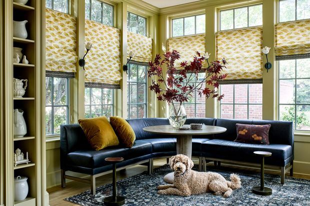 A bright, traditionally detailed sitting room or living room with extensive multi-pane bay-window glazing, patterned roller blinds, olive-green painted walls and trim, built-in display shelving, and a dark L-shaped leather sofa. Mustard and patterned cushions, a patterned area rug, small round side tables, a central vase of branches, and a dog complete the inviting seating area.