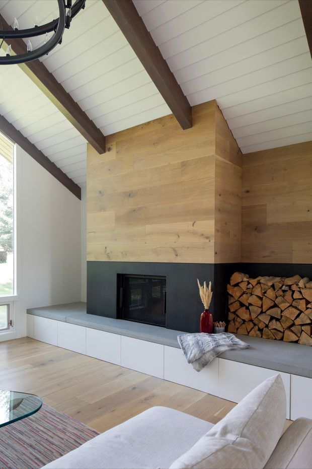 Bright contemporary living room featuring a double-height pitched ceiling with exposed dark timber beams, white V-groove ceiling boards, and a large timber-clad fireplace wall. The fireplace has a dark stone or metal surround, integrated white built-in cabinetry and bench seating, and an adjacent open firewood storage niche. A light neutral sofa, textured throw, rug, and modern black chandelier complete the warm, refined interior.
