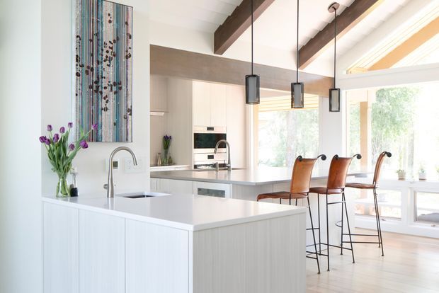 Bright contemporary kitchen with a large white island, integrated sink and tapware, pale cabinetry, stainless-steel appliances, and a secondary raised counter with leather bar stools. The space features exposed dark timber ceiling beams, pendant lights, extensive glazing, and a colourful vertical artwork.