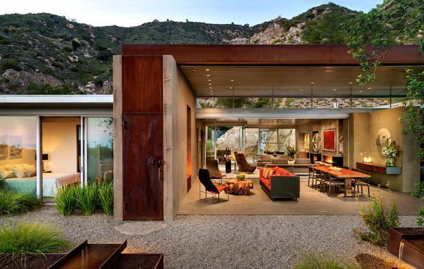 A contemporary single-storey residence integrated into a rugged rocky hillside. The exterior features a broad open-plan living pavilion with a deep rusted steel roof and framed openings, revealing a warm living and dining area with timber ceilings, extensive glazing, colourful seating, and artwork. A bedroom wing is visible at left, while gravel landscaping and native planting surround the house.