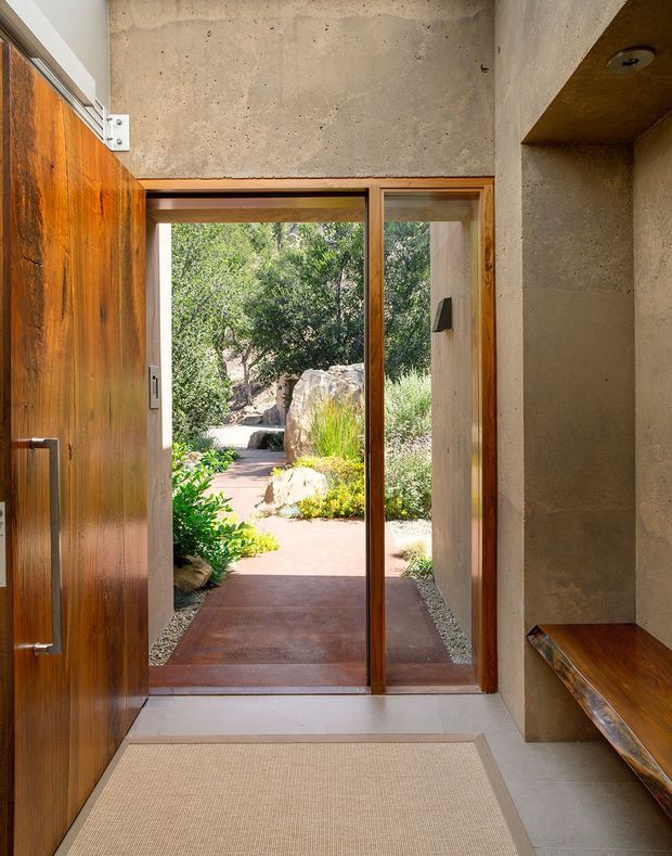A warm, minimalist entryway with a large solid timber pivot door, timber-framed glazed doorway, exposed textured concrete walls, and a built-in timber bench. The view opens directly onto a landscaped garden courtyard with rocks, planting, and a paved path.