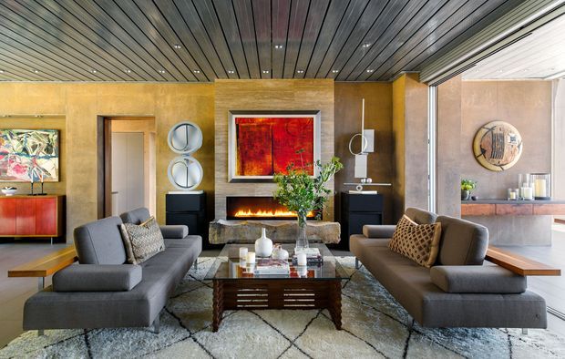 A sophisticated contemporary living room with two large grey sofas facing a stone-clad fireplace and wall-mounted artwork. A dark timber slatted ceiling, warm textured wall finishes, built-in cabinetry, glass-fronted openings, and a polished coffee table create a refined, layered interior. A dining or kitchen area is visible beyond the opening on the right.