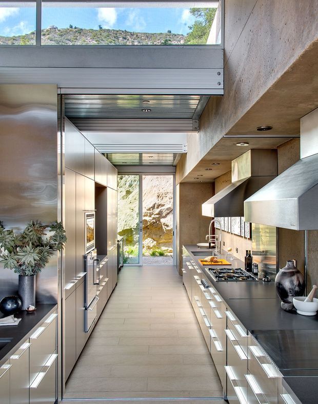 A narrow, galley-style contemporary kitchen with full-height stainless-steel and metallic cabinetry on both sides, dark stone-like benchtops, integrated appliances, and a gas cooktop beneath a large angular metal rangehood. The kitchen has a long central circulation path leading to glazed doors and a rocky outdoor setting. A linear rooflight or clerestory glazing brings in daylight, while exposed concrete-toned wall and ceiling surfaces add an industrial architectural character.