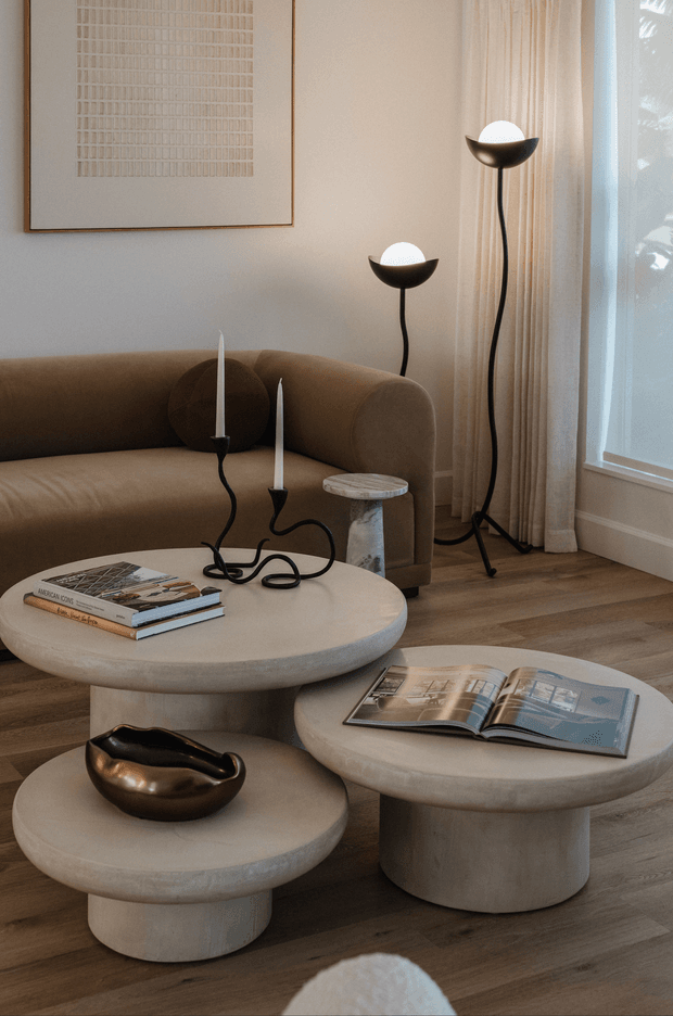 A warm, refined living room vignette with a rounded brown sofa, sculptural nesting coffee tables, black globe floor lamps, candles, books, and a metallic decorative bowl. Neutral plaster walls, sheer curtains, timber flooring, and a large abstract textured artwork create a soft contemporary setting.