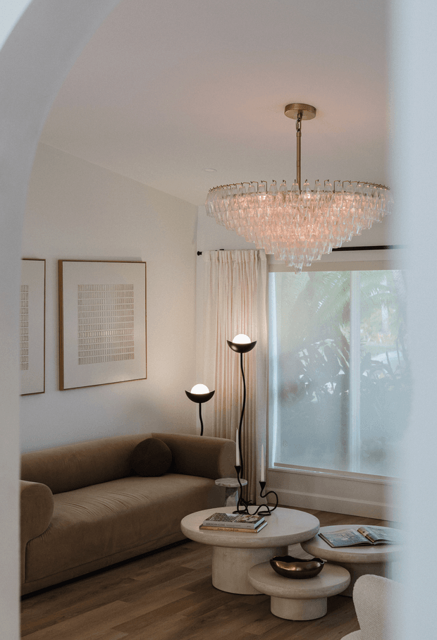 A refined living room viewed through a broad arched opening, featuring a curved taupe sofa, layered round travertine-style coffee tables, sculptural black floor lamps, framed minimalist artwork, full-height curtains, and a large cascading crystal chandelier. Soft daylight filters through the window, creating a warm, elegant atmosphere.