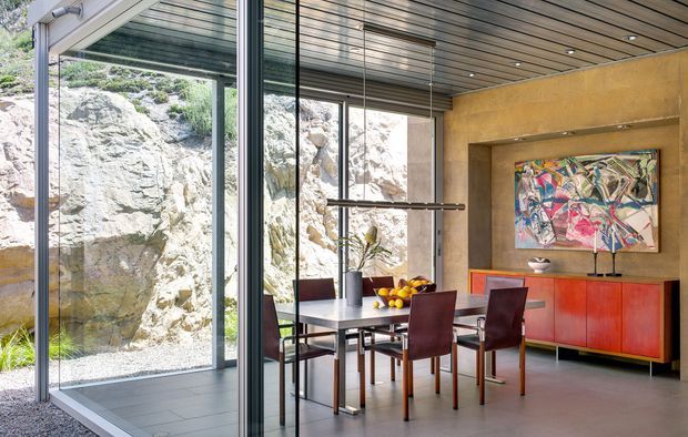Contemporary dining area with floor-to-ceiling glazing overlooking a rocky landscape. The room features a dark dining table with burgundy upholstered chairs, a vivid red-orange sideboard, timber-slat ceiling, warm metallic wall finish, pendant lighting, and colourful abstract artwork.