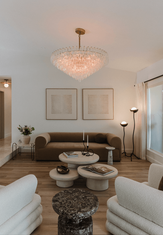 Warm, refined living room with a brown upholstered sofa, sculptural cream nesting coffee tables, textured white lounge chairs, a dark stone pedestal table, framed neutral artwork, floor lamps, curtains and a large crystal chandelier. Timber flooring and soft neutral finishes create an elegant, inviting atmosphere.