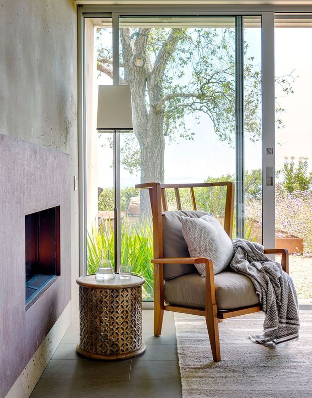 A bright, intimate living area with a mid-century-inspired timber armchair, neutral upholstery, patterned throw, and small decorative side table positioned beside a floor-to-ceiling glazed door or window. A built-in fireplace with a dark inset opening is set into a textured pale wall. The space opens visually to a leafy outdoor setting and distant water or sky, with soft natural light and warm, restrained materials.