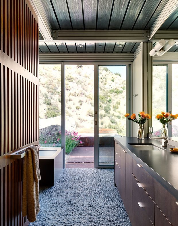 A compact contemporary kitchen with dark wood cabinetry, a long dark benchtop, integrated sink and chrome tapware, and full-height glazed doors opening to a landscaped outdoor area. The room features a distinctive pebble-textured floor, a vertically slatted timber feature wall, exposed dark ceiling panels with recessed lighting, and a large mirror reflecting the kitchen and windows.