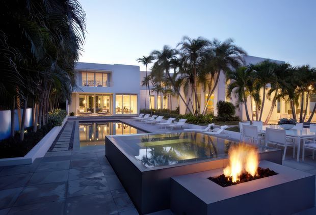 A luxurious modern courtyard and pool terrace at dusk, featuring a rectilinear multi-level reflecting pool, swimming pool, outdoor fire feature, dark stone paving, palm landscaping, and white contemporary architecture with expansive glazing. Outdoor dining and lounge furniture are arranged around the water features.