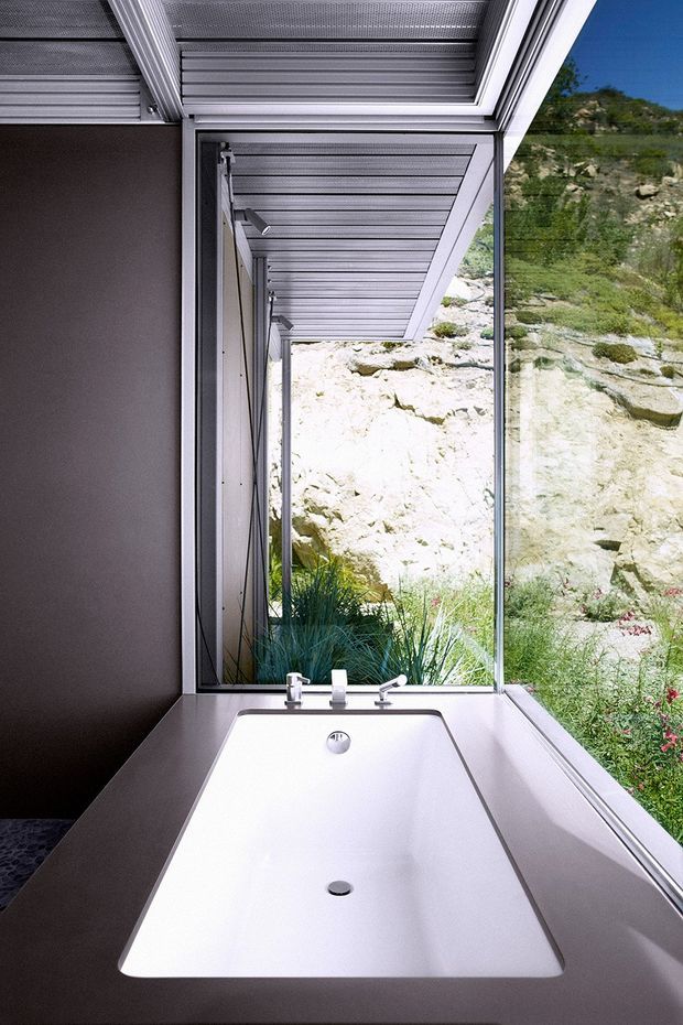 A modern minimalist bathroom featuring a freestanding white soaking bathtub set into a dark taupe or grey surround. Large floor-to-ceiling glazing beside the bath overlooks a rocky, landscaped hillside, while a projecting slatted metal canopy creates strong linear texture overhead. The space is calm, restrained and highly connected to the natural landscape.