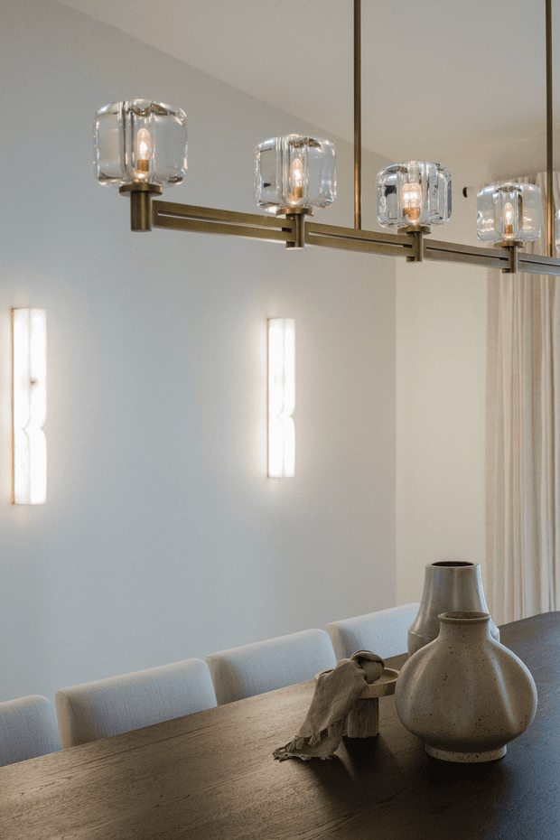 A refined dining area featuring a long dark timber dining table, upholstered light dining chairs, sculptural ceramic vessels, and a linear multi-light pendant with clear glass shades and warm brass-toned metalwork. Pale walls, vertical wall sconces, and sheer curtains create a calm, softly lit atmosphere.