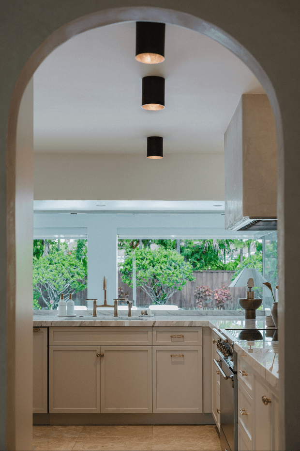 A warm, refined kitchen viewed through a large rounded archway. The room features cream painted cabinetry with brass hardware, a pale stone benchtop, integrated appliances, a central sink beneath wide windows, and three black cylindrical pendant lights. Large glazed openings provide views to a leafy garden, while the polished stone floor and soft neutral finishes create a classic yet contemporary atmosphere.