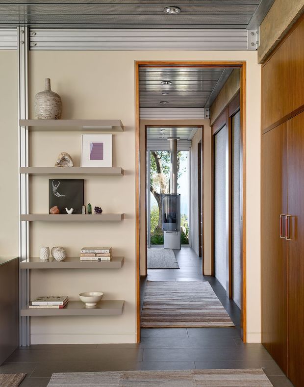 A refined interior hallway viewed toward a long glazed corridor, with built-in open display shelving on the left, tall timber cabinetry on the right, dark tiled flooring, warm wood door frames, and a distinctive corrugated metal ceiling with recessed downlights. A freestanding fireplace or heater is visible deeper in the corridor, with a calm neutral palette and layered rugs.
