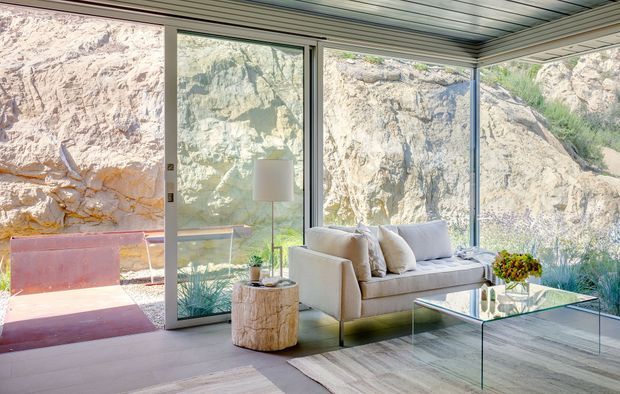 Bright contemporary living room with floor-to-ceiling glazing overlooking a rugged rocky landscape. The room features a neutral sofa, glass coffee table, pale timber-look flooring, a natural log side table, and a slatted ceiling, creating a clean indoor-outdoor connection.