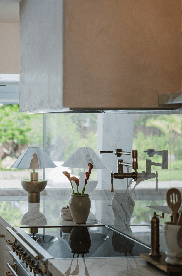 Close-up view of an elegant kitchen with a large marble island and reflective glass induction cooktop, brass-toned range controls, sculptural pendant-style lamps, and a prominent aged metal range hood. A marble backsplash and exposed industrial-style plumbing fixtures sit in front of large windows overlooking greenery. Decorative vessels and cooking utensils add warmth to the refined, layered composition.