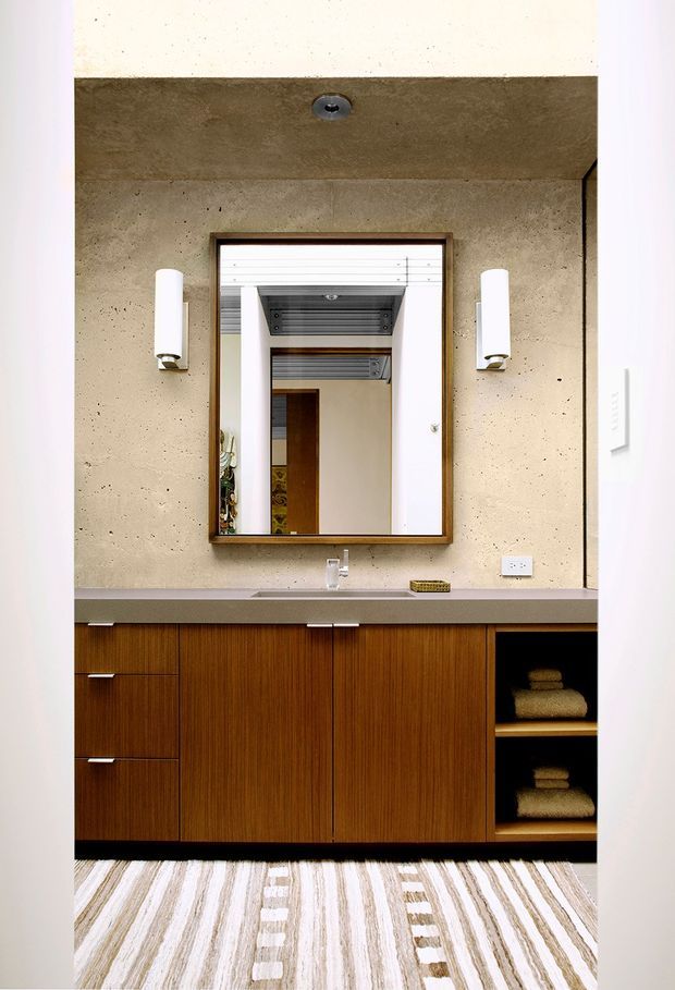 A refined bathroom vanity featuring a long dark timber built-in cabinet with open shelving for folded towels, a slim grey benchtop with an integrated basin, and a large framed mirror. The walls are finished in textured concrete, complemented by cylindrical wall sconces and a striped neutral area rug. The mirror reflects additional bathroom cabinetry and a linear ceiling vent, reinforcing the contemporary architectural character.
