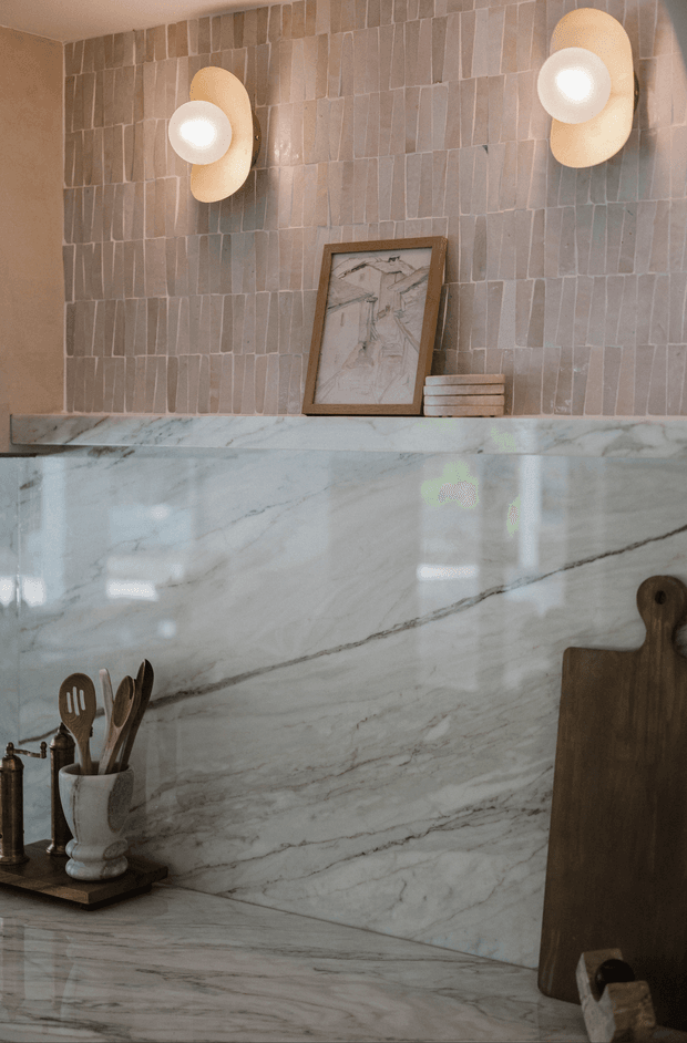 Close-up view of a refined kitchen or kitchenette fit-out featuring a dramatic veined white natural-stone benchtop and full-height splashback, a warm taupe-grey vertically arranged stone-tile wall, sculptural wall sconces, timber chopping boards, and a small still-life arrangement of timber utensils and framed artwork. The palette is warm, muted, and tactile with a softly luxurious feel.
