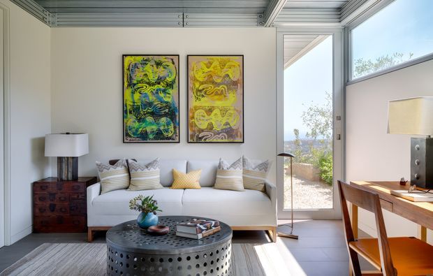 Bright, compact contemporary sitting room with a white sofa, patterned cushions, colourful abstract artwork, a perforated round metal coffee table, timber sideboard, and a timber desk with chairs. Large glazed door and windows bring in abundant daylight and overlook an outdoor area. The ceiling features exposed linear metal decking or panelling, creating an industrial architectural accent.