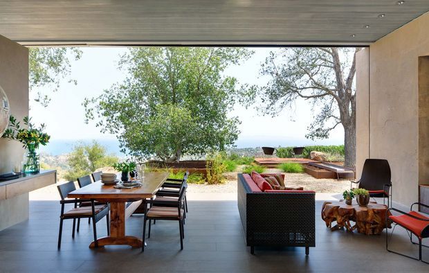 Open-plan covered outdoor living and dining area with a contemporary concrete and timber-lined structure, opening onto a landscaped terrace and distant landscape. The space contains a timber dining table with chairs, an outdoor sofa, lounge chairs, built-in cabinetry, and sculptural wooden side tables.
