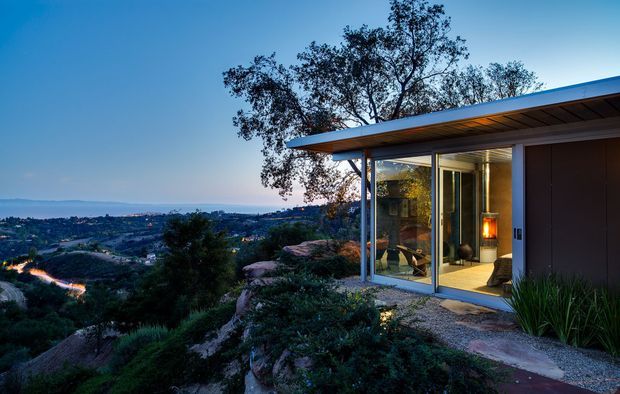 Twilight exterior view of a modern single-storey pavilion set into a rocky, landscaped hillside. The building has a thin flat roof with exposed timber soffit, dark wall cladding, slender metal-framed floor-to-ceiling glazing, and a glazed corner opening onto a narrow paved path. The interior is visible through the windows, while the surrounding valley landscape and distant suburban settlement form the background.