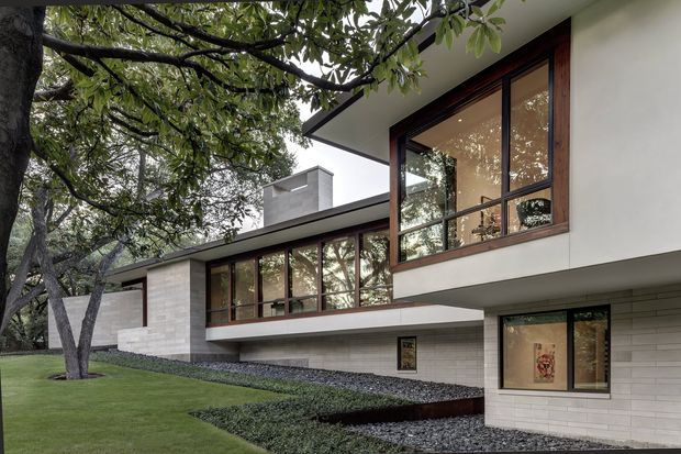 A low-slung modern residence set within a landscaped garden, viewed from outside. The house features strong horizontal volumes, projecting cantilevered upper sections, extensive dark timber-framed glazing, pale masonry cladding, and a flat roof. Mature trees frame the facade, while gravel beds and clipped planting surround the building.