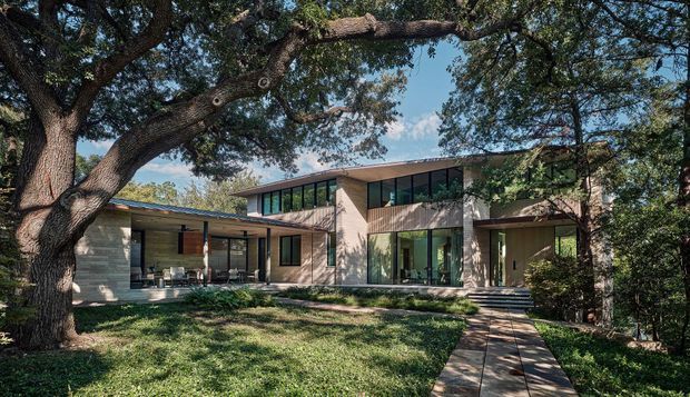 A contemporary multi-wing residence set within a mature, wooded landscape. The home features low-pitched roof forms, extensive floor-to-ceiling glazing, pale masonry or rendered walls, warm timber soffits, and a paved path leading through the lawn to the entrance. Large established trees frame the facade and create a strong connection between the interior and garden.