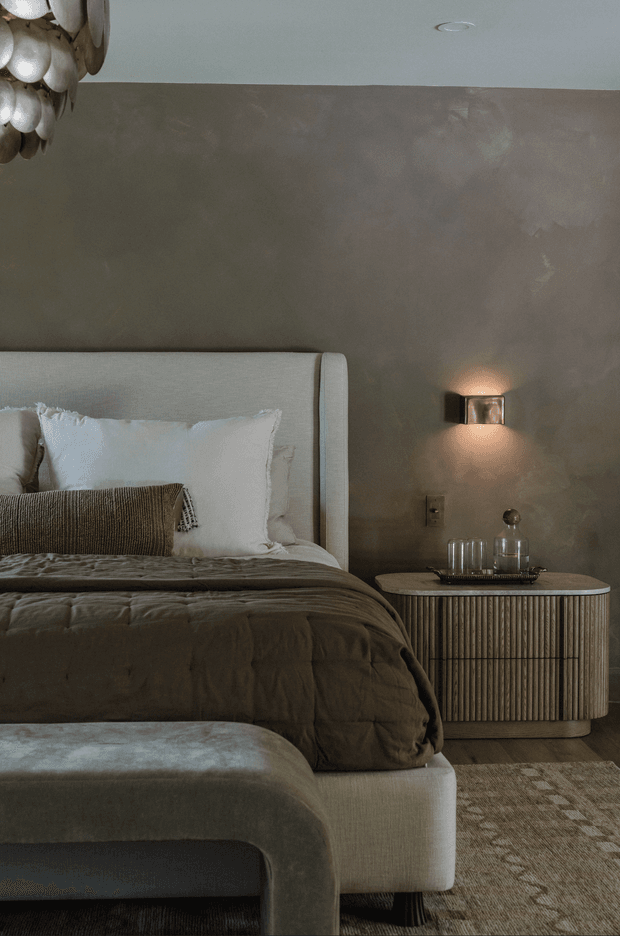 A moody, refined bedroom interior featuring a fully upholstered bed with a tall rounded headboard, layered neutral bedding and pillows, a fluted bedside cabinet, warm wall lighting, textured taupe plaster walls, and a softly patterned neutral rug. A sculptural pendant is partially visible at the upper left.