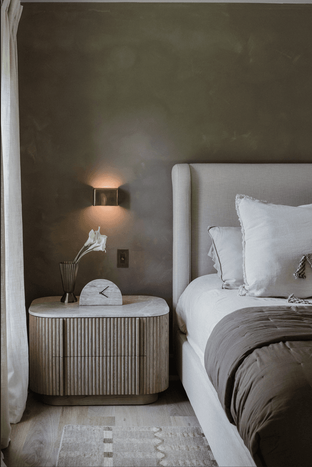 A softly lit, refined bedroom vignette featuring a tall upholstered bed with white bedding and a taupe throw, beside a fluted wood bedside cabinet with rounded edges. The dark taupe wall has a minimalist brass-toned wall sconce, while sheer curtains, pale timber flooring, and a muted patterned rug create a warm, calm atmosphere.
