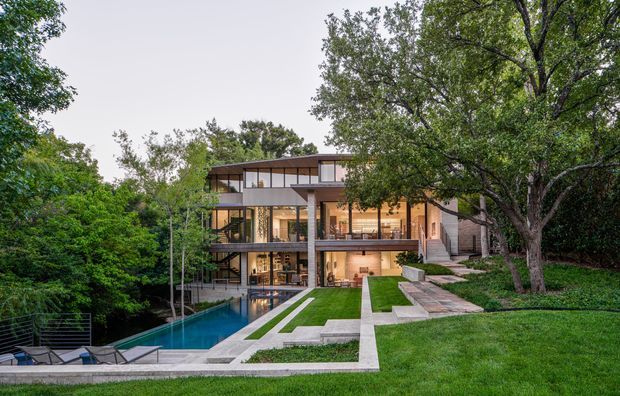 A contemporary multi-level residence set into a lush, sloping suburban landscape. The home features extensive floor-to-ceiling glazing, warm timber-toned interiors, projecting balconies, exposed concrete structural elements, and a long rectangular swimming pool with pale stone paving and manicured lawn terraces in the foreground.