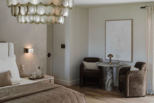 A refined bedroom sitting area with a partially visible upholstered bed, a tufted neutral bedspread, sculptural shell-like pendant lighting, and a small seating arrangement beside a veined stone-top table. The room has warm neutral walls, timber flooring, soft curtains, a wall sconce, and understated contemporary furnishings.