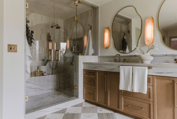 A refined contemporary bathroom with a large glass-enclosed walk-in shower, marble-look tiled walls and floor, a long double vanity with warm wood cabinetry and a pale stone benchtop, sculptural asymmetrical mirrors, and integrated warm decorative lighting. Brass-toned fixtures and hardware add a subtle luxe accent.