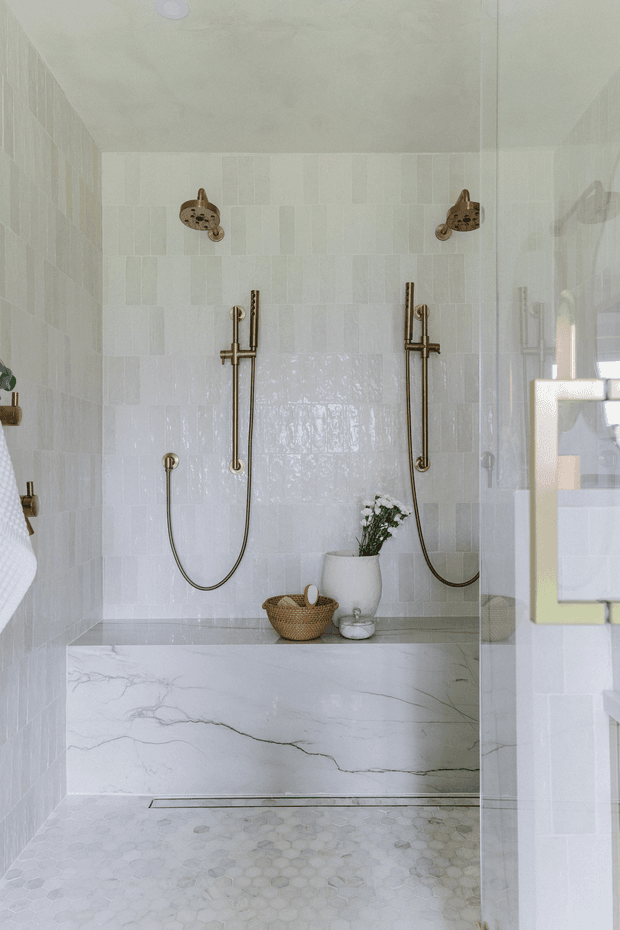 Bright walk-in shower with white and pale grey rectangular wall tiles, a marble-look stone bench, and small hexagonal floor tiles. Twin antique brass shower fixtures and handheld sprays are mounted on the rear wall, with a glass enclosure and a few simple natural accessories creating a refined, spa-like atmosphere.