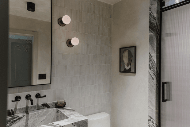 A refined contemporary bathroom viewed through a large wall mirror. The space features a sculptural marble vanity with wall-mounted dark metal taps, a white toilet, pale vertically arranged textured wall tiles, warm plastered walls, round globe wall lights, and a dark-framed shower enclosure with a ribbed translucent glass door and marble-clad surround.