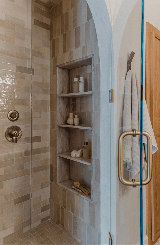 A close-up view of a tiled walk-in shower with warm beige and taupe rectangular wall tiles, a darker tiled recessed niche with multiple shelves, brushed brass shower controls, and a frameless glass shower door with a large brass handle. A white textured towel hangs beside a natural timber door.