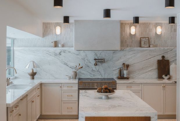A refined light-toned kitchen with extensive veined natural-stone surfaces, including a large island and full-height backsplash. Pale shaker-style cabinetry lines the walls, with a central range, brass-toned hardware, integrated sink area, decorative cutting boards, and pendant lights. The space is bright and balanced, with a polished contemporary-classic feel.