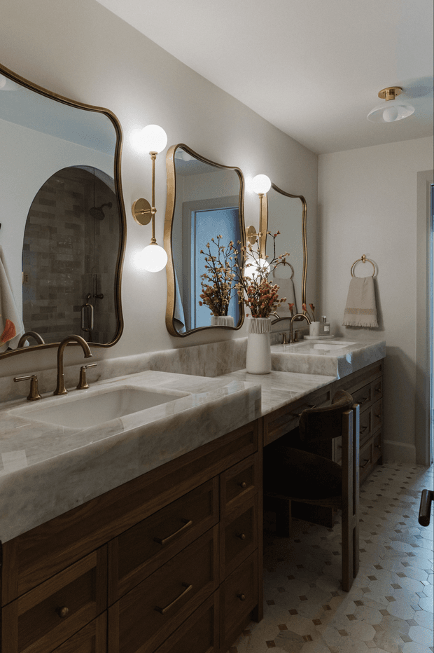 A warm, refined bathroom with a long double vanity featuring dark timber cabinetry and a pale marble benchtop with integrated basins. Three organically shaped bronze-framed mirrors are paired with globe wall sconces. A tiled shower is visible in the reflection, while patterned floor tiles, brass-toned tapware, a small upholstered stool, and floral branches complete the space.