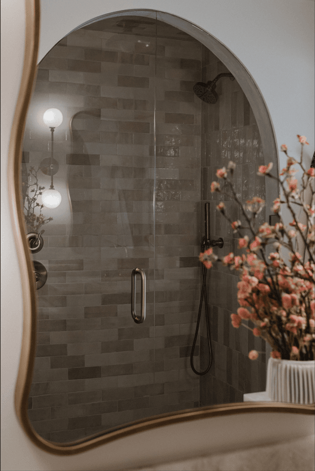 A close-up interior view of a refined bathroom shower enclosure, primarily seen through a large rounded-edge mirror. The walk-in shower has grey-brown rectangular wall tiles in a staggered layout, dark metal shower fittings, and a frameless glass door with a vertical handle. A softly lit vanity area and a ribbed white planter with flowering branches appear in the foreground, creating a warm, elegant atmosphere.