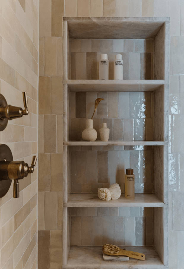 Close-up view of a built-in recessed shower niche with multiple shelves, set within a tiled bathroom wall. The niche has warm beige and cream rectangular tiles with varied tonal texture, paired with aged brass shower controls at the left edge. White toiletry bottles, small ceramic vessels, a flower, folded cloth, and a wooden brush provide restrained styling.