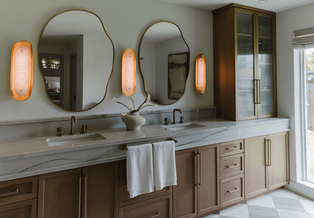 A refined double-vanity bathroom with a long pale veined stone countertop, two integrated sinks, sculptural dark-framed mirrors, warm orange wall sconces, and extensive two-tone timber cabinetry. A tall glass-front storage cabinet and full-height window add vertical emphasis and natural light.